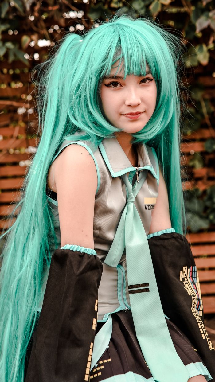 A young woman cosplaying as a popular character with vibrant turquoise hair outdoors.